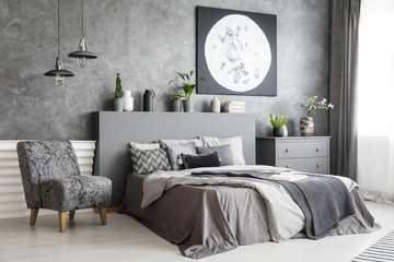 Armchair next to bed in grey bedroom interior with moon poster on concrete wall. Real photo
