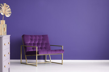 Wide armchair, chest of drawers and golden decorations set on a purple, empty wall. Place your...