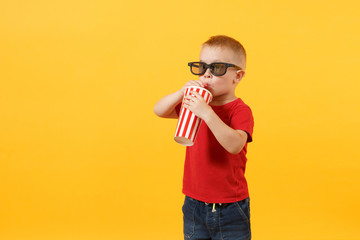 Little cute kid baby boy 3-4 years old in red t-shirt, 3d imax cinema glasses holding plastic glass of soda, drinking cola isolated on yellow background. Kids childhood lifestyle concept. Copy space.