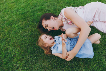 Laughing woman in light dress and little cute child baby girl lie on green grass lawn tickle, play...