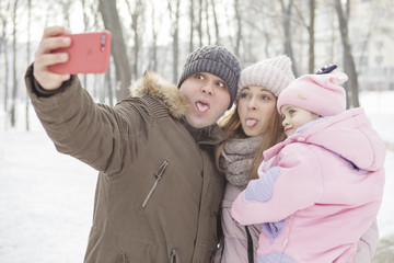 Happy loving caucasian family of mother father and daughter play, having fun in winter snowy park. They photograph themselves (make selfie) on a phone