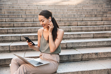 Image of beautiful smiling woman 20s wearing casual summer outfit and bluetooth earphone, sitting on street stairs and holding smartphone while using silver laptop