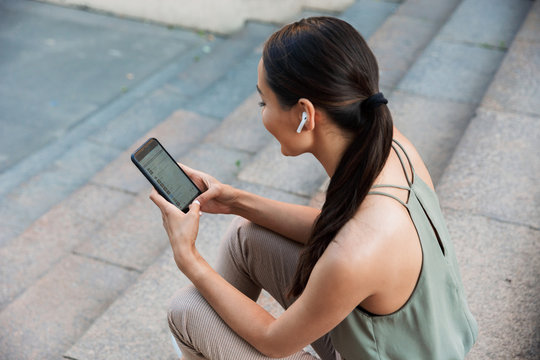 Image from top of brunette young woman wearing casual outfit, sitting on street stairs on summer day while listening to music via bluetooth earpod and mobile phone