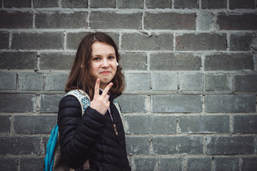 Schoolgirl with backpack stands against gray brick wall with artificial emotion with copy space. Not real smile of depressed teen in faded tones. Young girl with headphones in cloudy weather.