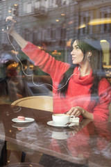 Cute brunette plus size woman working on a distance (freelance), sitting in cafe, drinking coffee and eating macaron. She wearing red sweater, listen music and make selfie on her phone