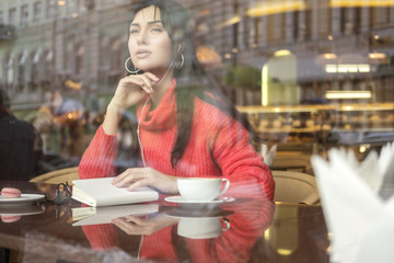 Cute brunette plus size woman working on a distance (freelance), sitting in cafe, drinking coffee and eating macaron. She wearing red sweater, listen music and typing messages on her phone