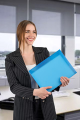 young successful woman in a strict dark suit with a blue file in hands looking aside and smiling in office