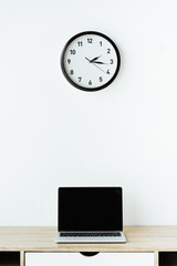 laptop on work desk under clock hanging on white wall