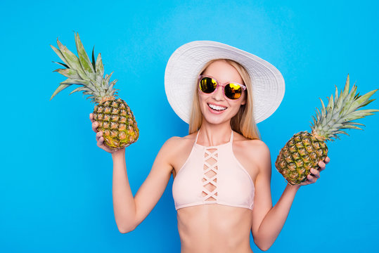 Portrait of cheerful pretty girl in pink bra summer eye glasses holding two fresh pineapples in hands looking at camera isolated on bright blue background