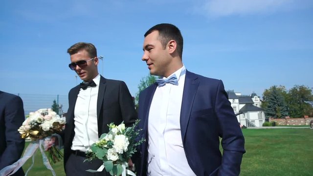 Handsome groom and his best men with wedding bouquets are walking along the garden and talking.