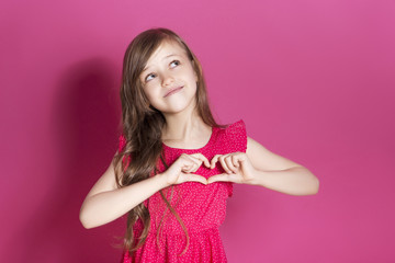 Little 8 years old girl make some emotional gesture with her hands on a pink neutral background. She has long brunette hair and wear red summer dress. Funny expression on her face