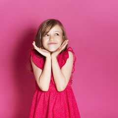 Little 8 years old girl make some emotional gesture with her hands on a pink neutral background. She has long brunette hair and wear red summer dress. Funny expression on her face