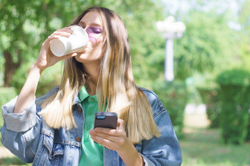 Attractive young girl is smiling and standing with a mug of coffee and a smartphone. Fashionable hipster model in trendy heart-shaped sunglasses. Concept of messaging and texting in the park