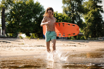 Slim long-haired beach lifeguard running in water with life-saving equipment
