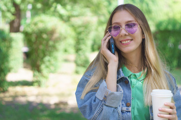 Naklejka premium Attractive young girl is smiling and standing with a mug of coffee and a smartphone. Fashionable hipster model in trendy heart-shaped sunglasses. Concept of messaging and texting in the park