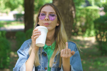 Beautiful young girl shows the middle finger. Fashionable hipster model in heart-shaped sunglasses. Funny concept of showing the frivolity of the situation and attitude