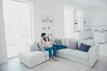 Full size portrait of attractive trendy couple covered with blanket having mugs with warm tea in hands, looking at each other, sitting in modern open space apartment
