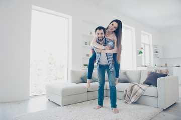 Full size low angle view portrait of crazy funny couple in denim outfits, bearded man carrying lover on back looking at camera, standing in modern open space white flat room with interior