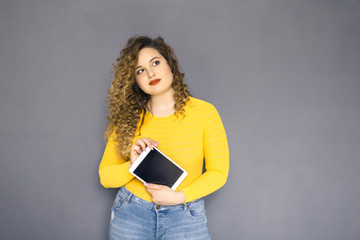 Cute brunette plus size woman with curly hair in yellow sweater and jeans standing on a neutral grey background. She holds tablet with her hands and look up thoughtfully. Space for text