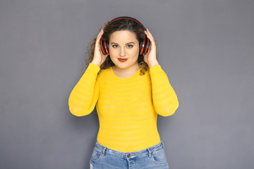 Cute brunette plus size woman with curly hair in yellow sweater and jeans standing on a neutral...