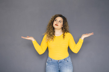 Cute brunette plus size woman with curly hair in yellow sweater and jeans standing on a neutral grey background. She is trying to decide what to choose. Holding hands and palms like plate for options