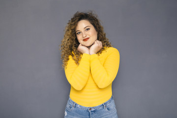 Cute brunette plus size woman with curly hair in yellow sweater and jeans standing on a neutral grey background. She was thinking, thoughtful emotion on her face. Copy space