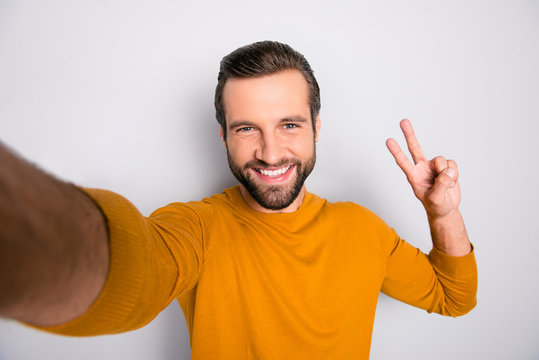 Self portrait of funky funny with toothy beaming smile bearded guy wearing yellow sweater taking making selfie demonstrating v-sign isolated on gray background