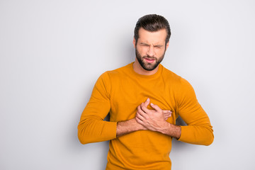 Broken heart concept. Portrait of grimacing sad upset nervous unhappy frustrated exhausted bearded man placing hands on chest suffering from pain in heart isolated on gray background copy-space