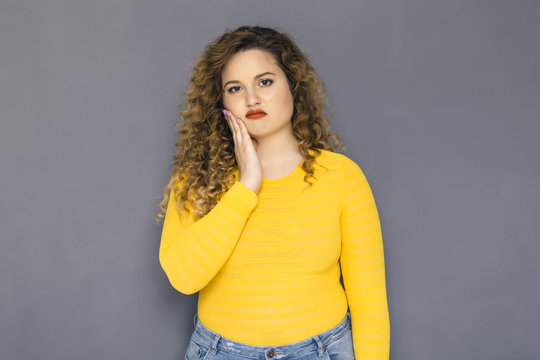 Cute brunette plus size woman with curly hair in yellow sweater and jeans standing on a neutral grey background. She has a thooth pain, holding her hand on her cheek
