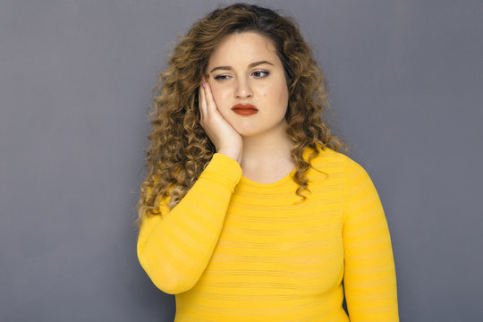 Cute brunette plus size woman with curly hair in yellow sweater and jeans standing on a neutral grey background. She has a thooth pain, holding her hand on her cheek