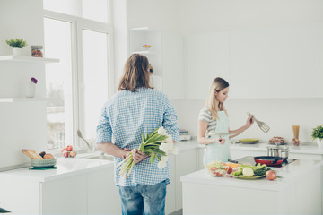 Portrait of handsome man holding bouquet of white tulips behind back in hands preparing surprise while housewife making meal dinner in modern kitchen with interior