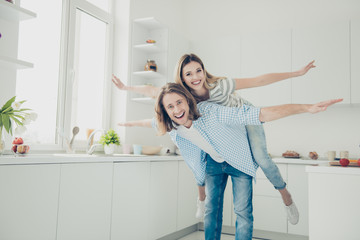 Portrait of positive fancy partners making airplane holding open hands to sides, handsome man carrying on back lover in modern white kitchen with interior