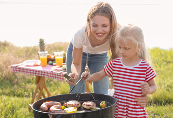 Young woman with daughter cooking meat, sausages and vegetables on barbecue grill outdoors
