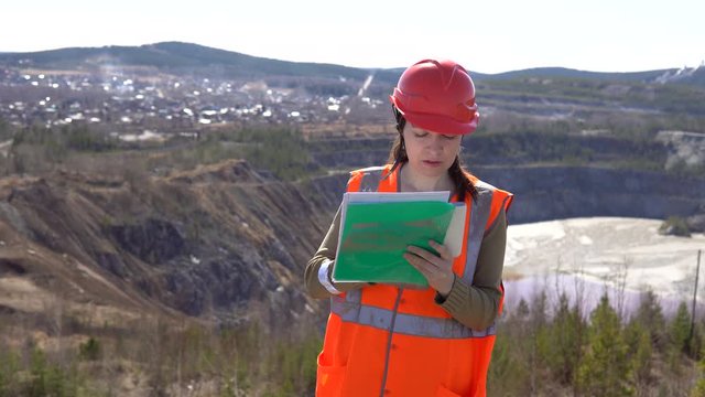 A young woman checks documents against the background of an open-air career on a sunny day. She is wearing an orange vest and a protective helmet.
