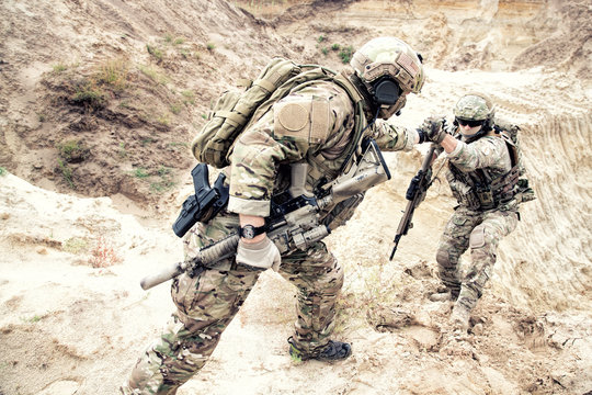 US ranger, modern infantryman, special reconnaissance team member or military scout in ammunition, armed with service rifle helping brother in arms to climb on sand dune. Army brotherhood and teamwork