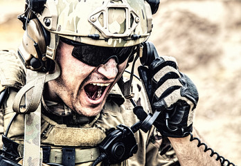 Special forces soldier, military communications operator or maintainer in helmet and glasses, screaming in radio during battle in desert. Calling up reinforcements, reporting situation on battlefield