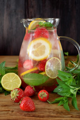 Lemonade with strawberry, lemon, mint, lime and ice cubes in a glass jug surrounded by the ingredients