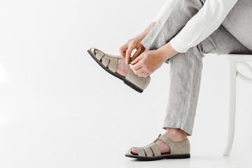 cropped image of male model in linen trousers sitting on chair and putting on sandals isolated on...