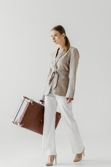 confident stylish female tourist in linen jacket holding vintage suitcase isolated on grey...