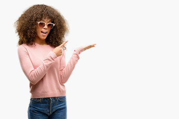 African american woman wearing pink sunglasses amazed and smiling to the camera while presenting with hand and pointing with finger.