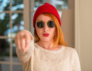 Stylish redhead woman wearing wool cap and sunglasses pointing with finger to the camera and to...