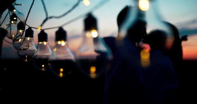 Romantic portrait of the hugging loving couple behind the string of lights during the sunset. 4k footage.