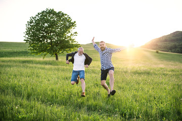 An adult hipster son and his senior father jumping in nature at sunset.