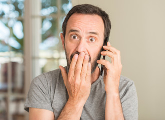 Middle age man using smartphone cover mouth with hand shocked with shame for mistake, expression of fear, scared in silence, secret concept