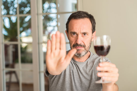 Middle age man drinking a glass of wine with open hand doing stop sign with serious and confident expression, defense gesture