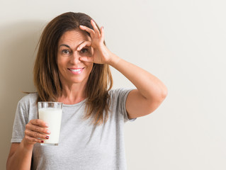 Middle age woman drinking a glass of fresh milk with happy face smiling doing ok sign with hand on eye looking through fingers