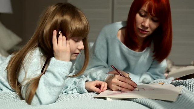 A daughter is drawing with her mother lying on the bed