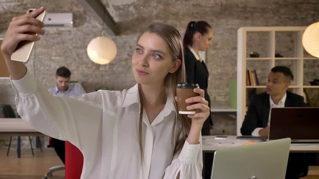 Young attractive businesswoman is making selfie with coffee on smartphone in office, her colleagues are networking with technologies, working concept, business concept, communication concept