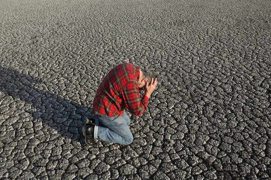 Desperate man kneeling and holding his head in his hands at dry cracked land after drought, natural disaster