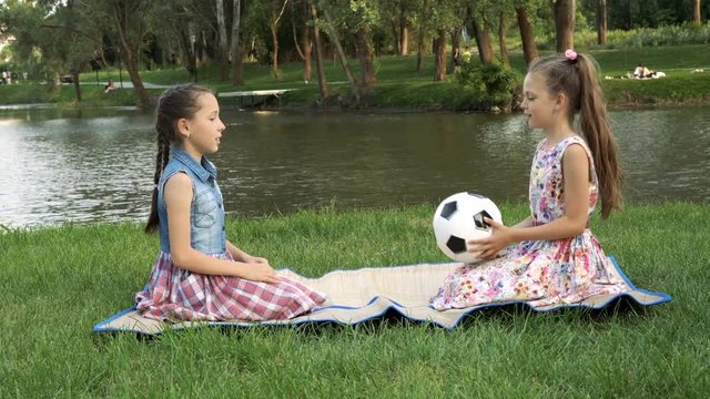 Two charming little girls sit at sunset in the Park on the lawn against the river and play with a soccer ball, throwing it to each other. 4K, 29.97 fps.